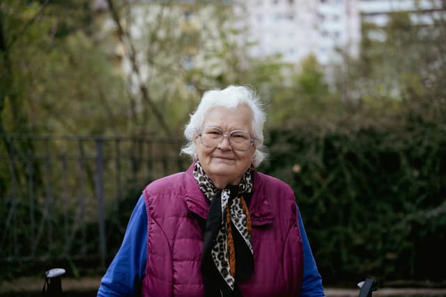 Smilende ældre kvinde med briller og lilla vest sidder udendørs i en park