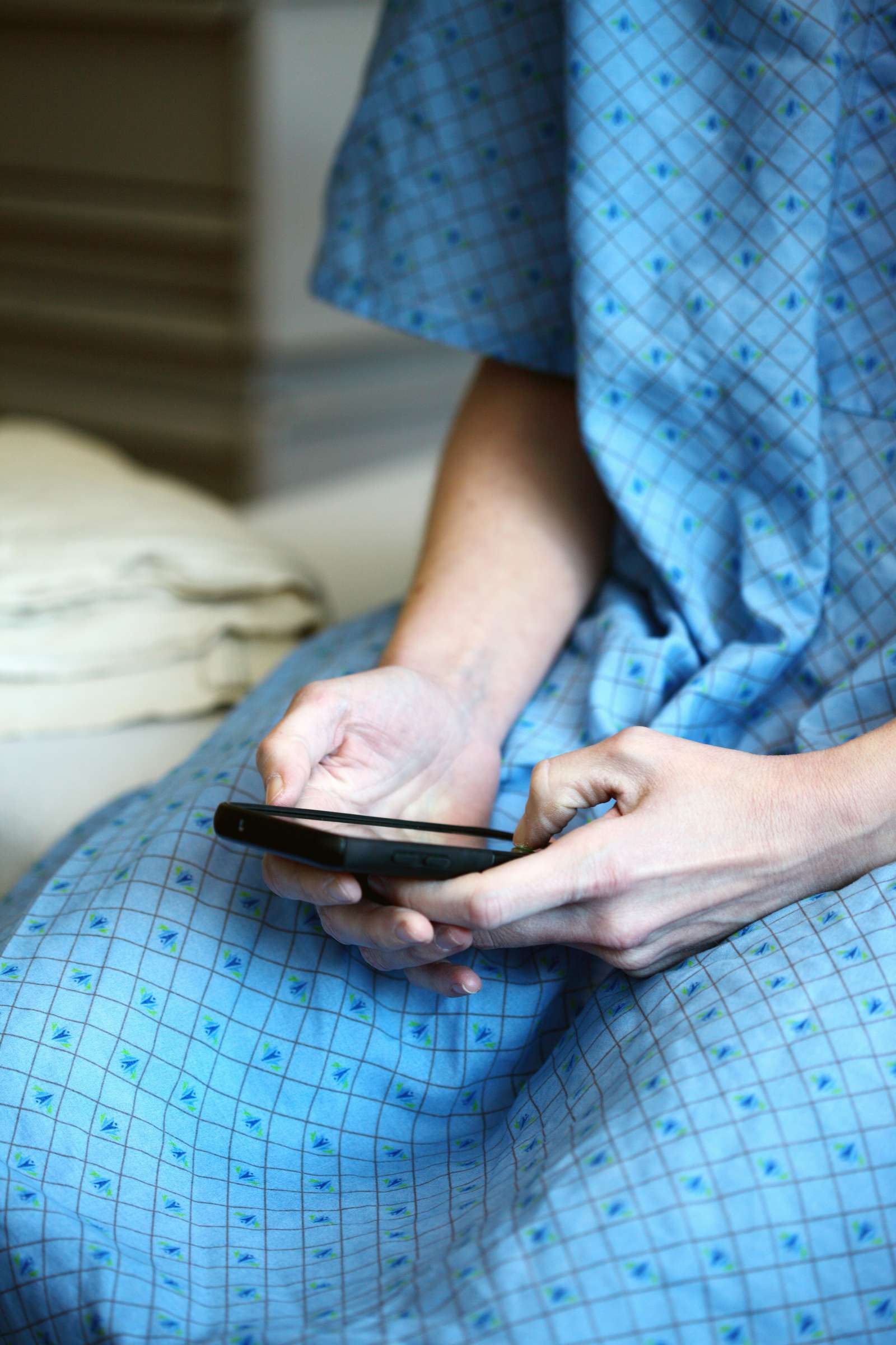 Patient i blå hospitalskjole sidder på briks og holder en smartphone i hænderne