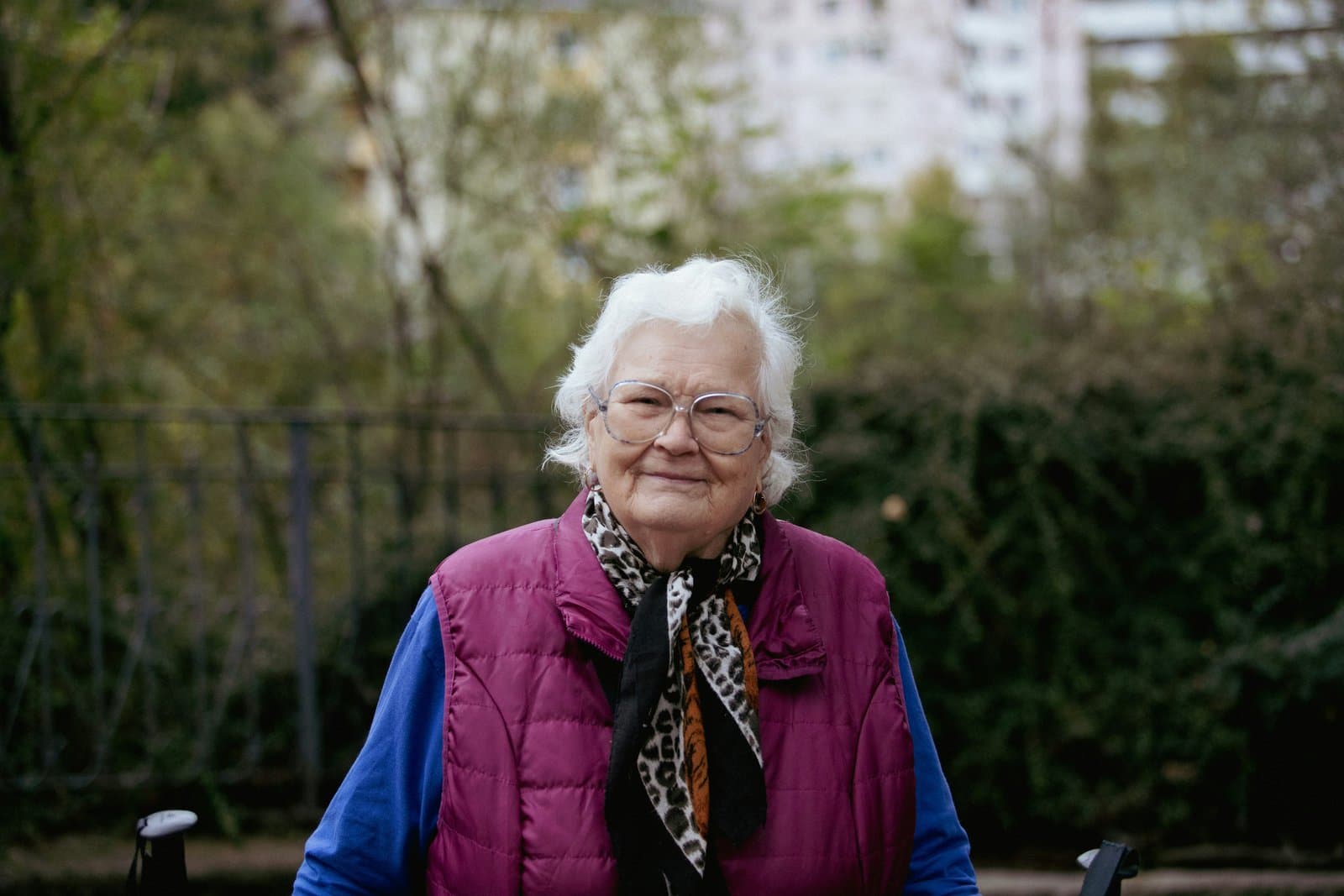 Smilende ældre kvinde med briller og lilla vest sidder udendørs i en park