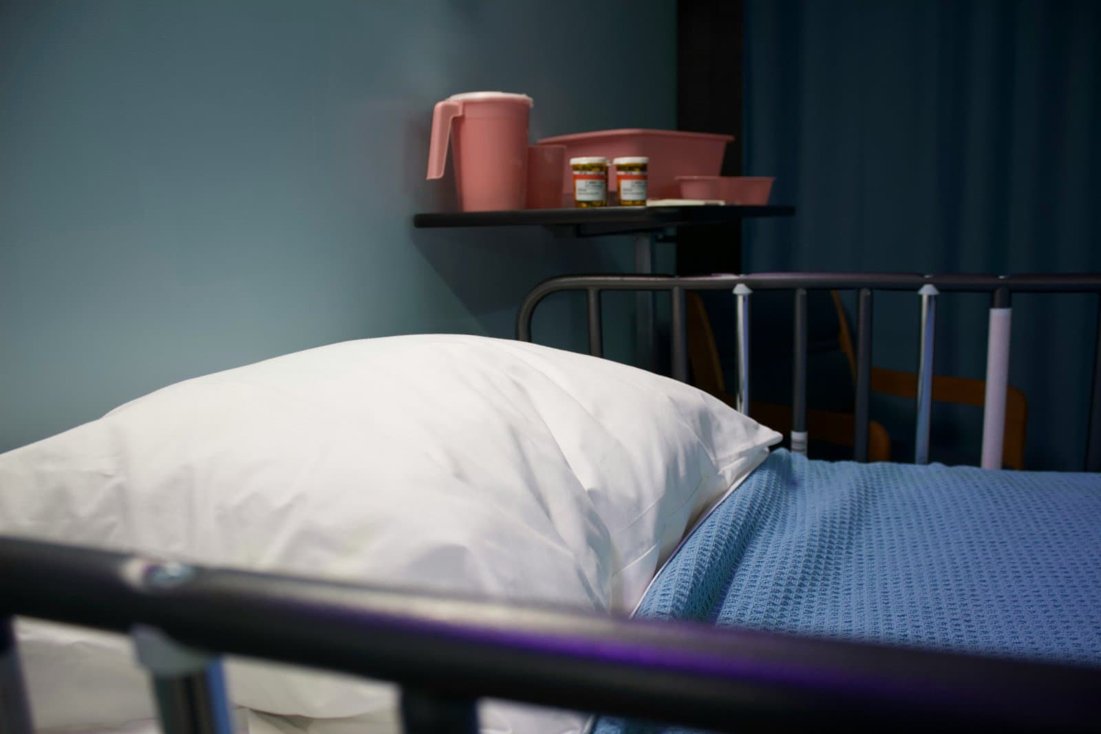 Tom hospitalsseng med hvid pude og blåt tæppe samt medicin på sengebord