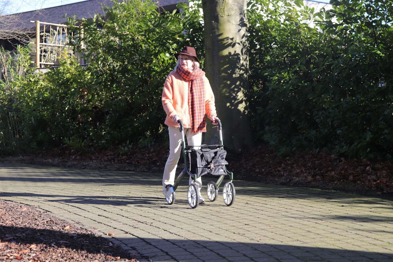 Ældre kvinde med hat og rollator går på en sti i en have en solrig dag