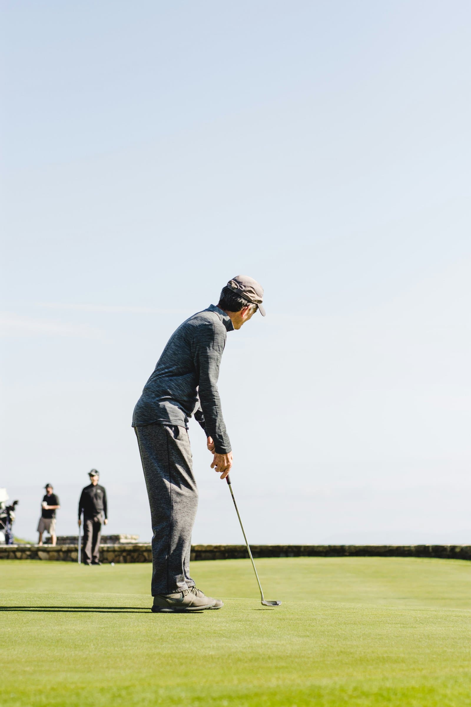 Ældre mand putter på golfbane en klar dag, to golfspillere i baggrunden